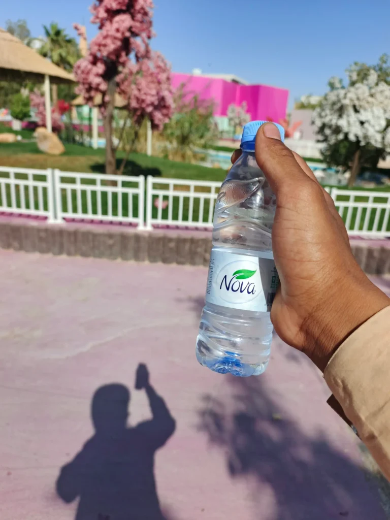 Hand holding Nova water bottle at Riyadh Zoo Pink Lake area showing hydration necessity during July summer visit